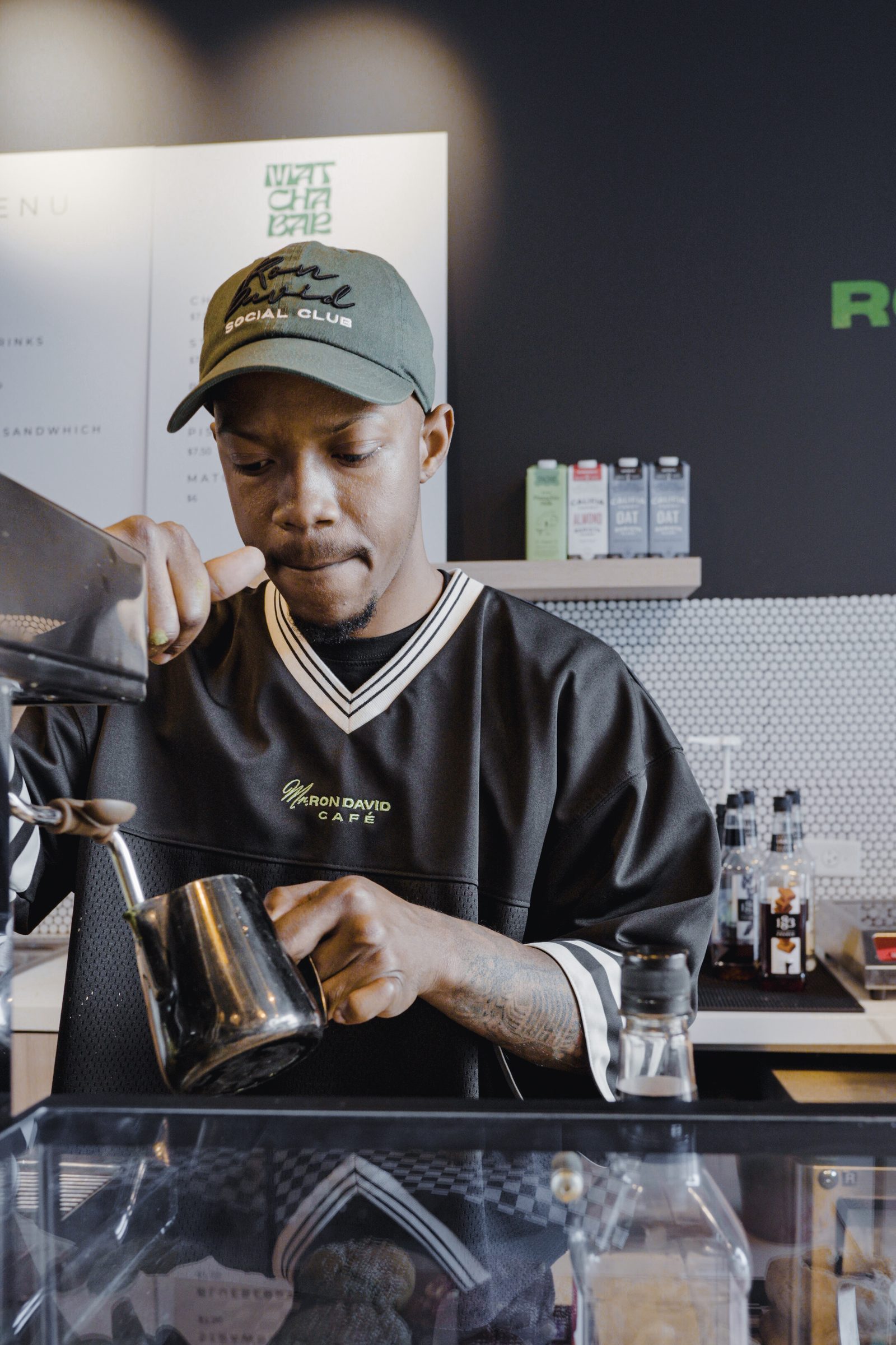 A barista in a Ron David cap and jersey pouring steamed milk at the coffee bar